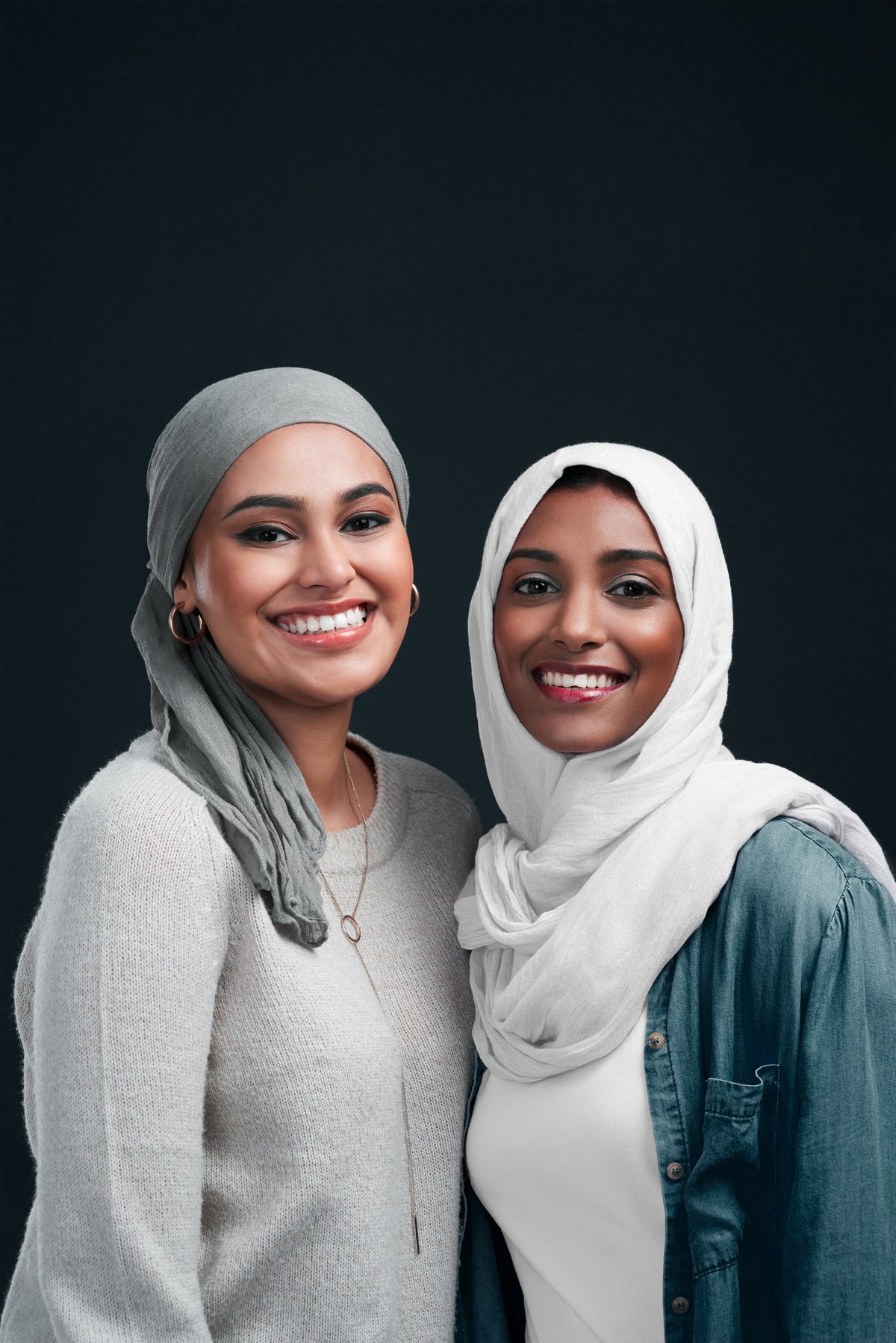 Two women in hijab standing close together against a black studio background