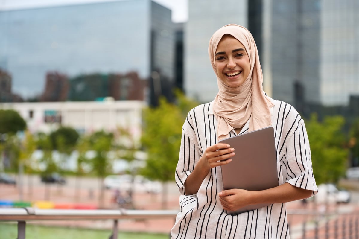 A smiling woman in hijab holding a laptop in the city