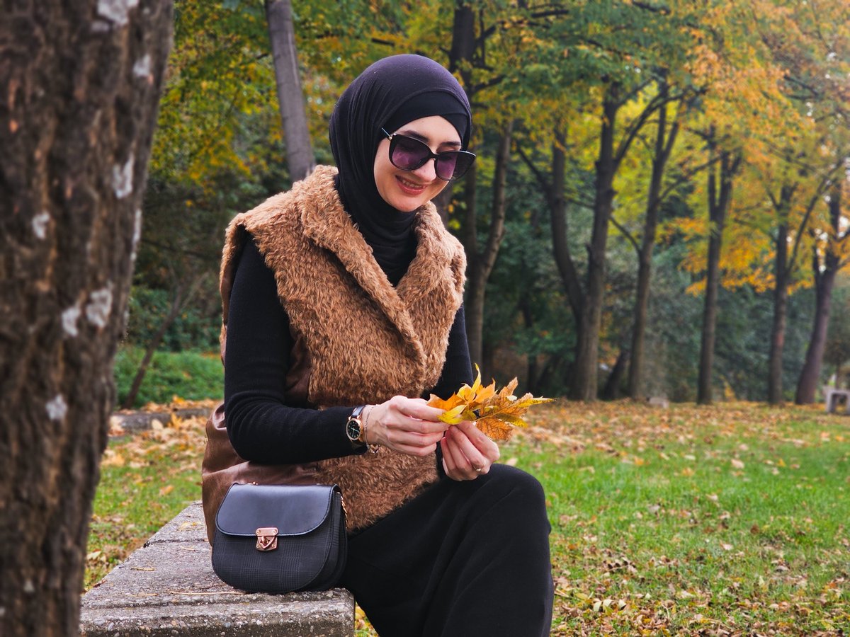 A stylish woman in hijab smiling while holding autumn leaves at a park