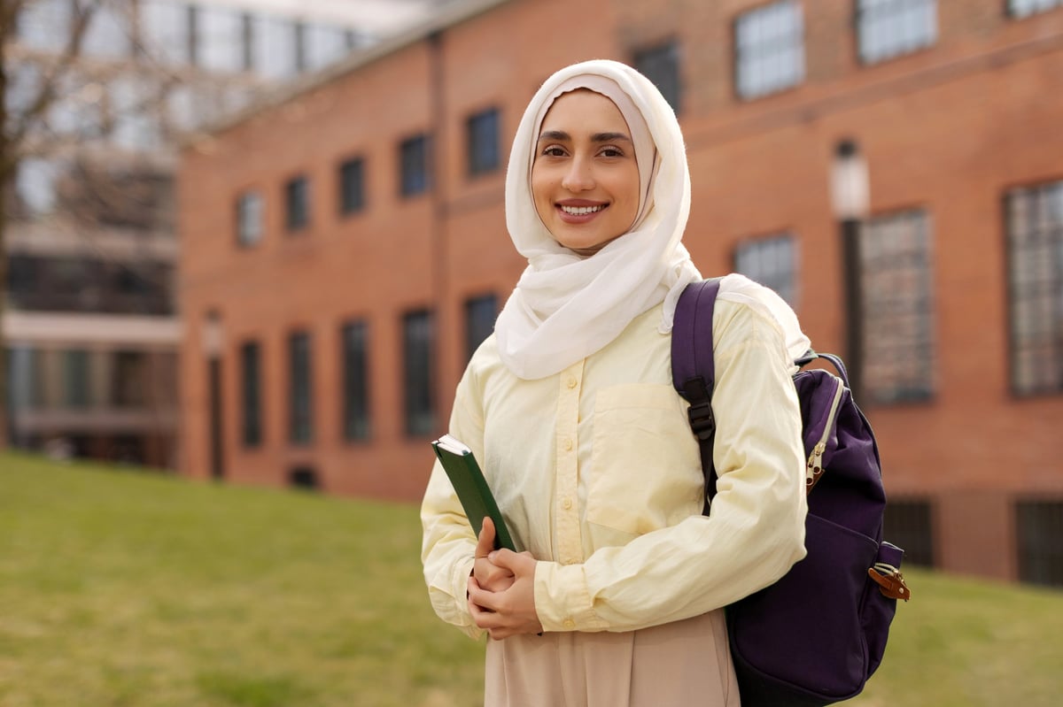 A happy student in hijab holding a book outside a university
